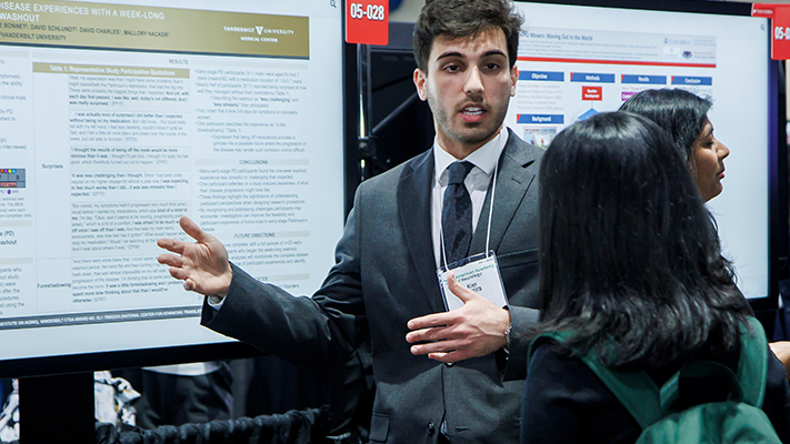 Man in a suit presenting a scientific research poster to an attendee at a medical conference.