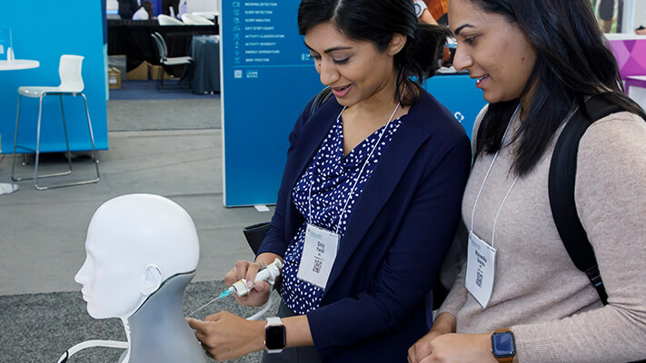 Two women interact with a medical simulation mannequin, demonstrating a technique at AAN conference exhibit.
