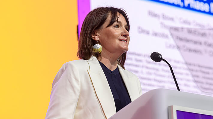 A woman in a white blazer speaks at a podium against a colorful AAN plenary background with text displayed on a screen.