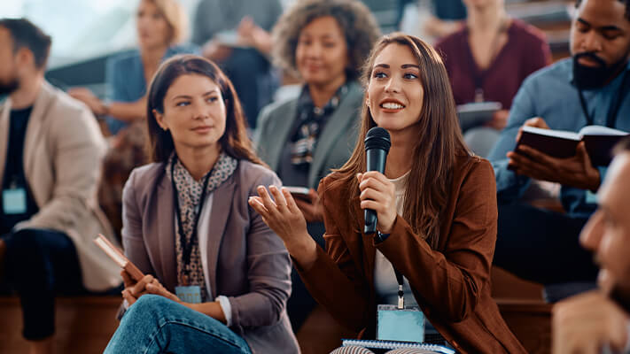 A woman speaks into a microphone during a conference, while an audience listens attentively from their seats.