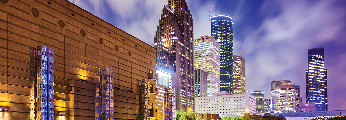 Night view of downtown Houston skyline with illuminated skyscrapers and modern buildings beside a large convention center exterior.