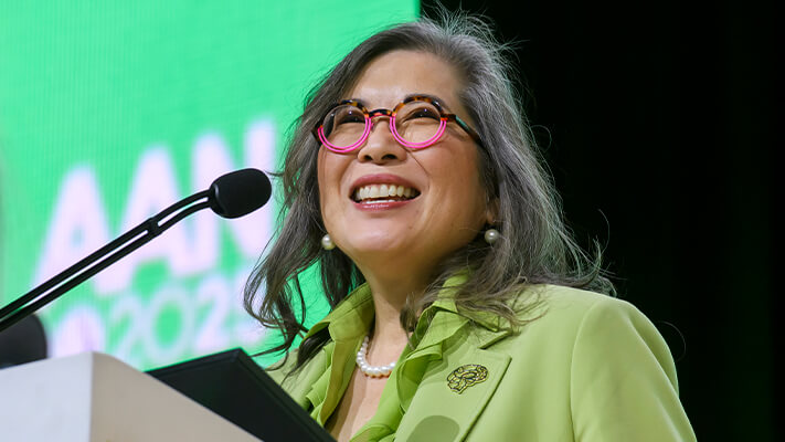 A woman in a green shirt speaks at a podium against a colorful ɫ background.