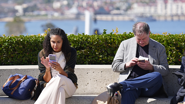 Two people sitting on a low concrete bench outdoors near a waterfront, each looking down at a smartphone, with bags beside them and a city skyline and water visible in the background.