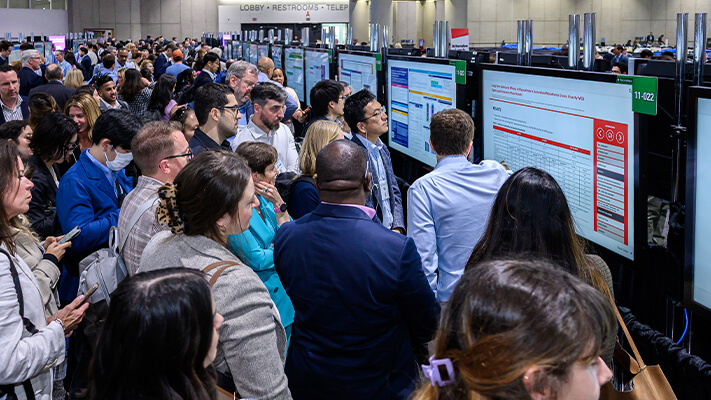 A large crowd gathers around digital displays in poster area, engaged in presentations and discussions.